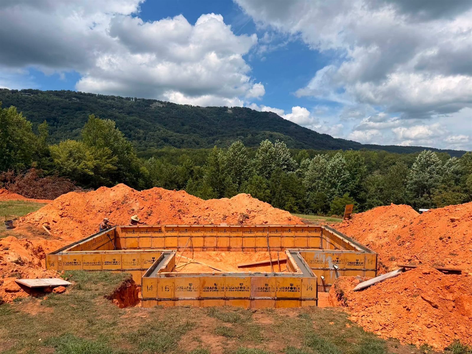 Crew setting steel wall forms for a pool with red clay spoil piles and a mountain behind