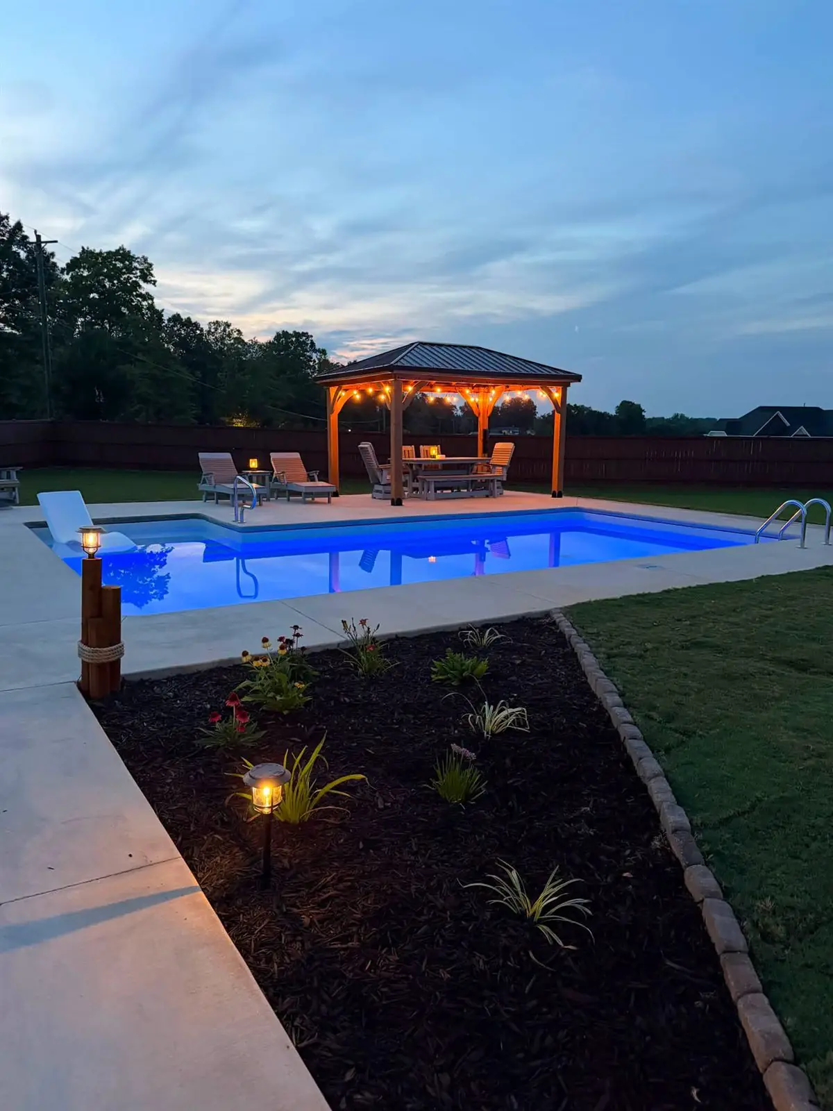 Pool lit blue at dusk with a lit gazebo, landscape lighting and a planted border