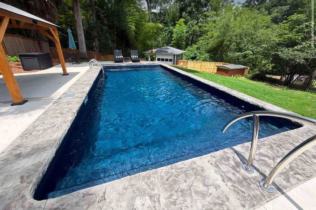 Rectangular pool with a deep navy liner and stamped stone coping beside a cedar pavilion and manicured lawn