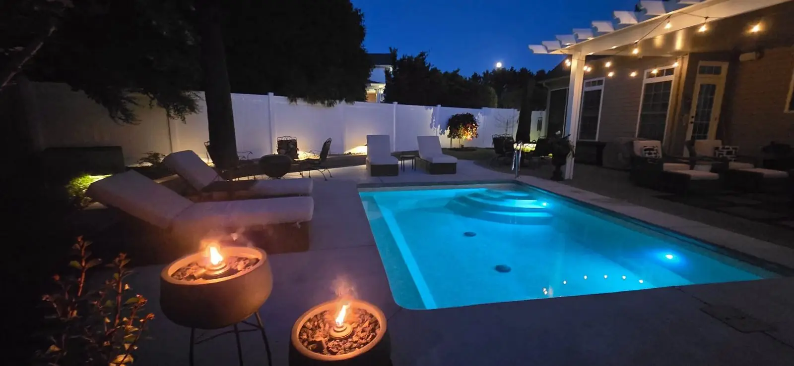 Backyard pool lit from within at night with fire bowls and string lights on the patio