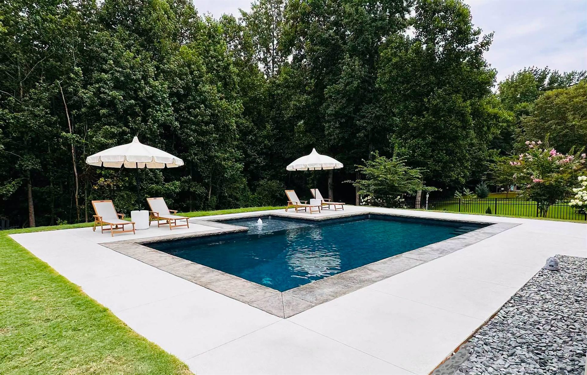 Rectangular in-ground pool with a tanning ledge, stone coping and shade umbrellas beside a wooded lot